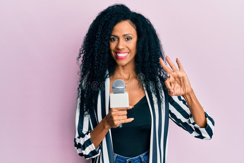 Middle age african american woman holding reporter microphone doing ok sign with fingers, smiling friendly gesturing excellent symbol. Middle age african american woman holding reporter microphone doing ok sign with fingers, smiling friendly gesturing excellent symbol