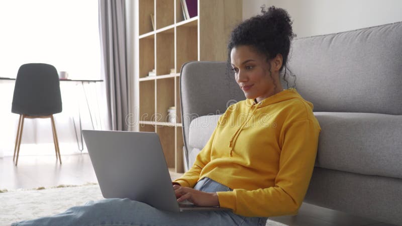 Afrikanisch-amerikanische Studentin sitzt zu Hause auf dem Boden und tippt auf Laptop.