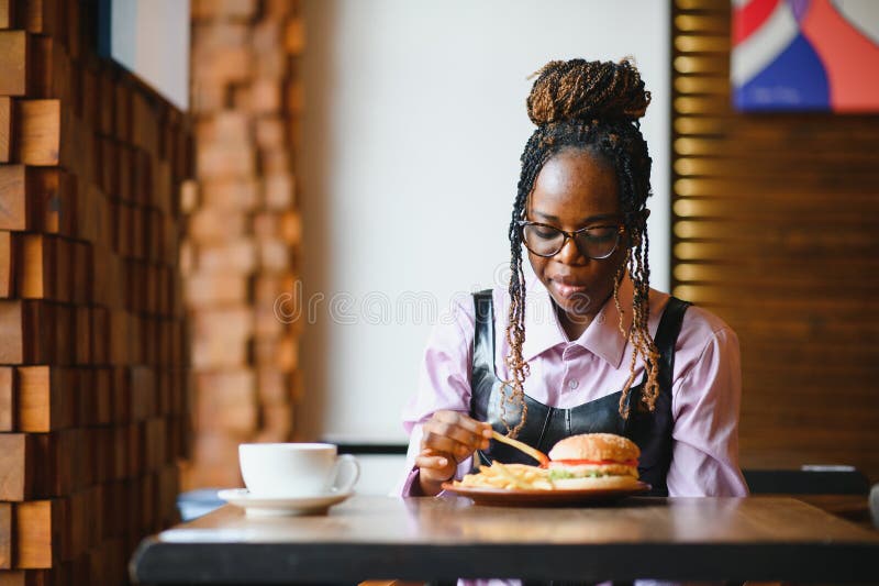 Afrikanische Frau mit afro Haar frisst einen leckeren klassischen Burger mit Pommes Frites. Cheat Meal stockfotos