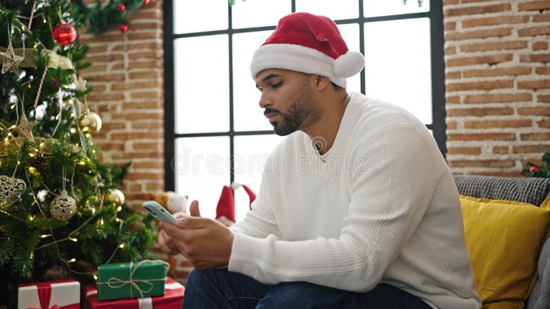 Afrikanischer Amerikaner, der Smartphone benutzt, Weihnachten feiert und zuhause verärgert aussieht stockbild