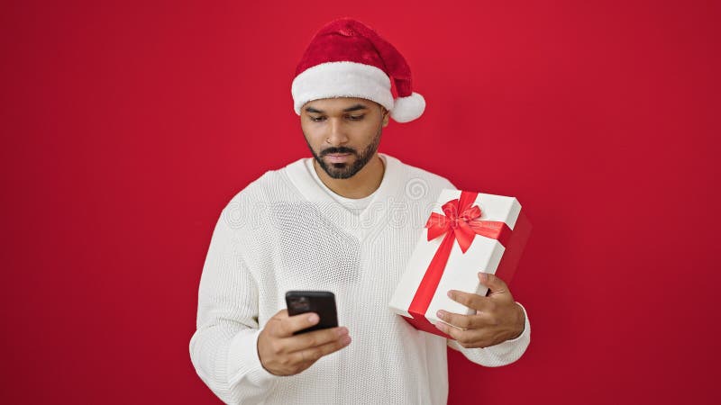 Afrikanischer amerikanischer Mann, der Weihnachtsgeschenk mit Smartphone über isolierten roten Hintergrund hält stockbild