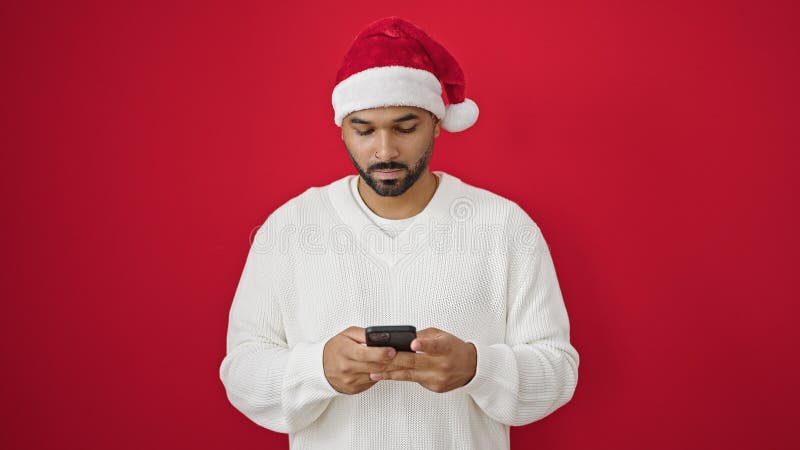 Afrikanischer amerikanischer Mann mit Weihnachtsmütze über isoliertem rotem Hintergrund mit Smartphone lizenzfreie stockfotografie