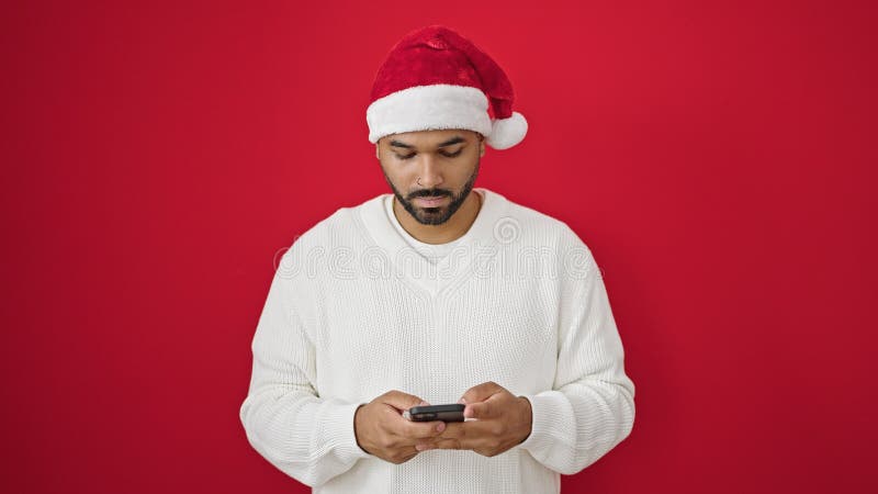 Afrikanischer amerikanischer Mann mit Weihnachtsmütze über isoliertem rotem Hintergrund mit Smartphone stockbild