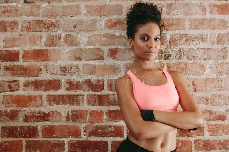 Portrait of young african woman in sports bra standing against brick wall with her arms crossed. Female fitness model in gym. Portrait of young african woman in sports bra standing against brick wall with her arms crossed. Female fitness model in gym.