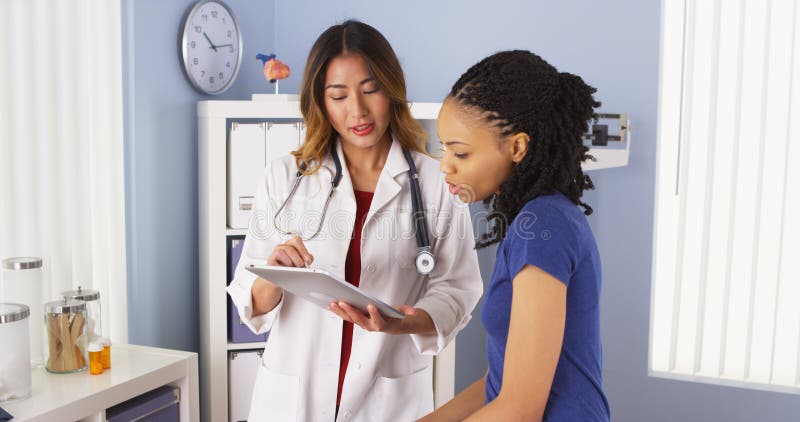 Afrikansk amerikanpatient som förklarar frågor till den asiatiska doktorn som använder minnestavlan royaltyfri foto