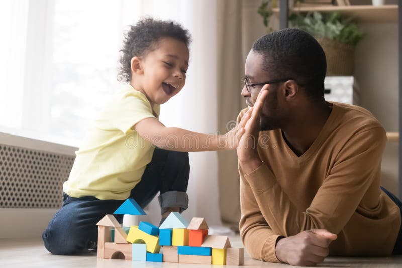 African American father in glasses with toddler son playing with colorful wooden constructor, giving five, adorable little child and black dad or babysitter having fun on warm floor at home. African American father in glasses with toddler son playing with colorful wooden constructor, giving five, adorable little child and black dad or babysitter having fun on warm floor at home