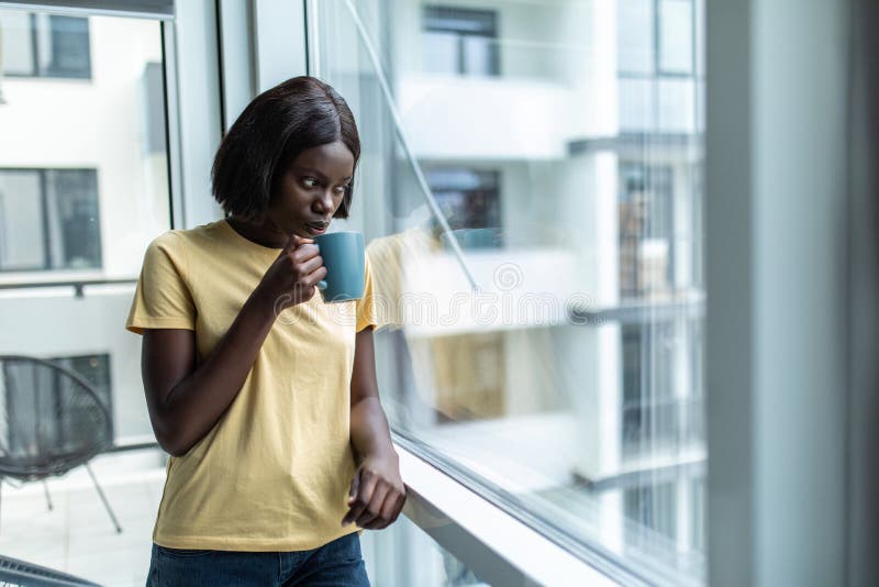 Afrikansk amerikansk kvinna i sitt vardagsrum som dricker en kopp kaffe och tittar på fönstret arkivbild