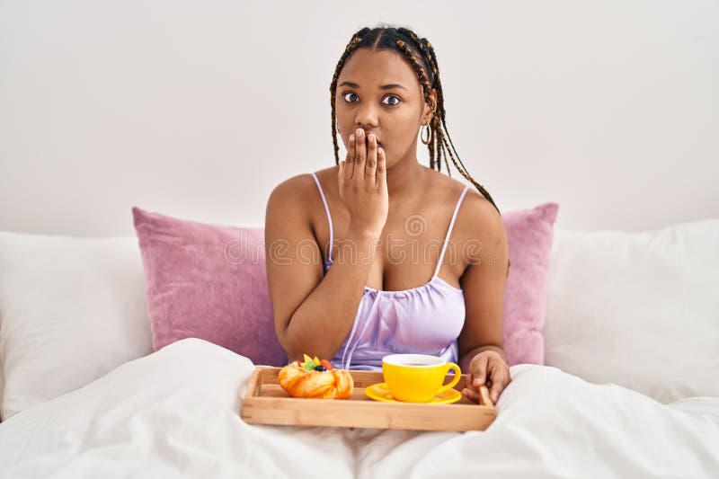 Afrikansk amerikansk kvinna med braida som håller i brickan med frukost i sängen som täcker munnen med hand chockad och rädd för. arkivfoto