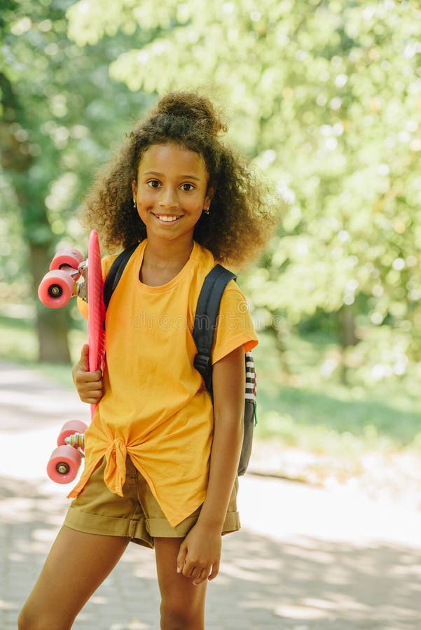 Afrikansk amerikansk skolflicka leende på kamera medan man håller skateboard royaltyfria bilder