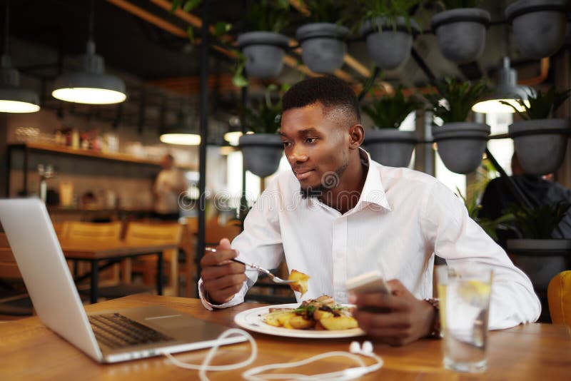 Handsome african man having dinner at stylish cafe, using and looking at laptop. Businessman in white shirt studding at restaurant, watching move, typing at call phone, writing message. Freelancer. Handsome african man having dinner at stylish cafe, using and looking at laptop. Businessman in white shirt studding at restaurant, watching move, typing at call phone, writing message. Freelancer.