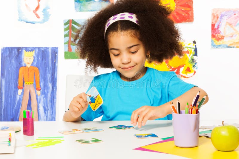 African girl putting matching cards during developmental game on table while sitting in playroom with wall behind full of children drawings. African girl putting matching cards during developmental game on table while sitting in playroom with wall behind full of children drawings