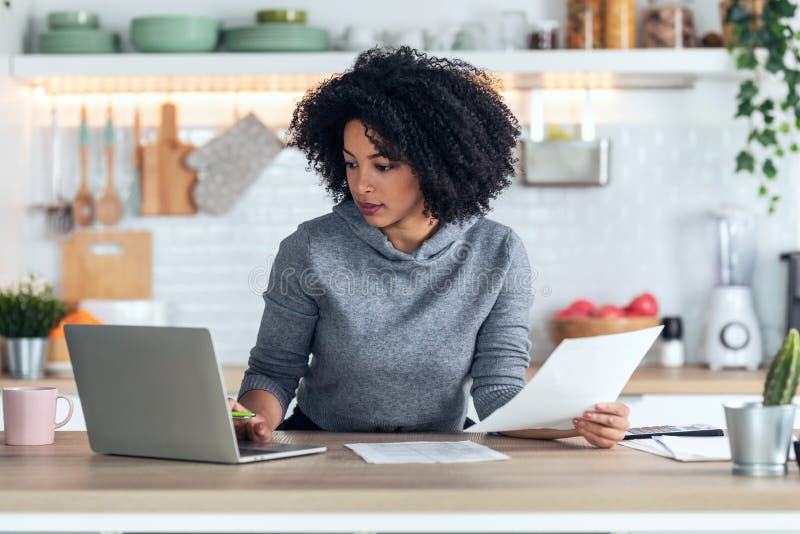 Afro business woman working with computer while consulting some invoices and documents in the kitchen at home