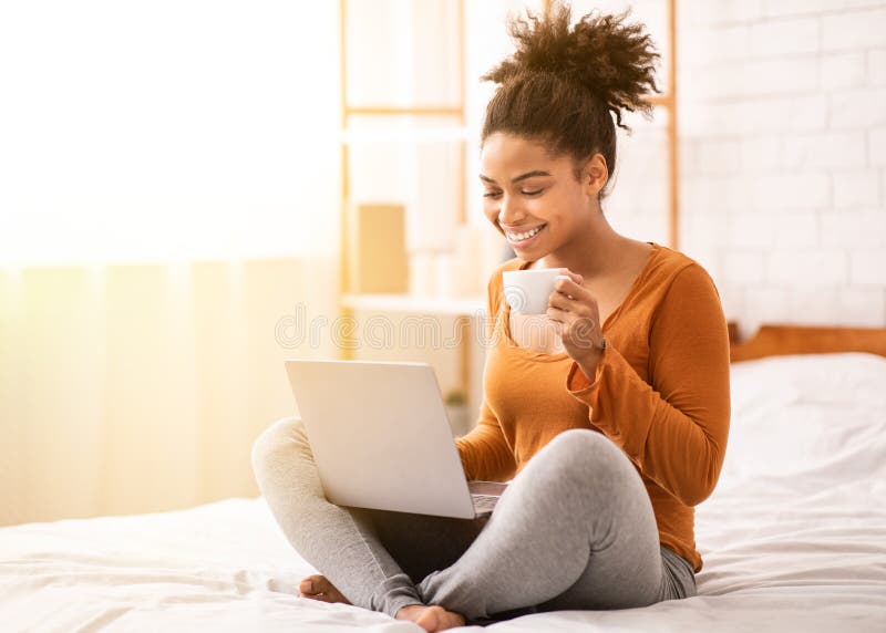 Afromujer sonriente que usa laptop sentada en el dormitorio imagen de archivo libre de regalías