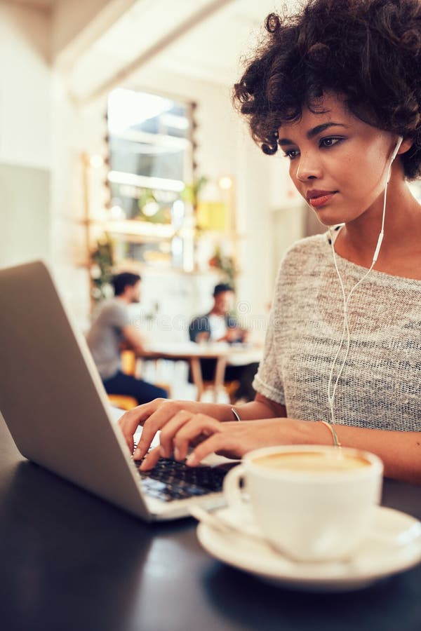 Portrait of attractive young woman with earphones working on laptop at a cafe. African female at a coffee shop surfing internet on laptop. Portrait of attractive young woman with earphones working on laptop at a cafe. African female at a coffee shop surfing internet on laptop.