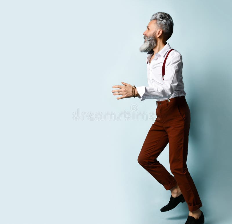Aged man in white shirt, brown pants and suspenders, black loafers. He runs looking back, raised his hand up, blue background. Gray-haired bearded man in a white royalty free stock images Aged man in white shirt, brown pants and suspenders, black loafers. He runs looking back, raised his hand up, blue background. Gray-haired bearded man in a white royalty free stock images