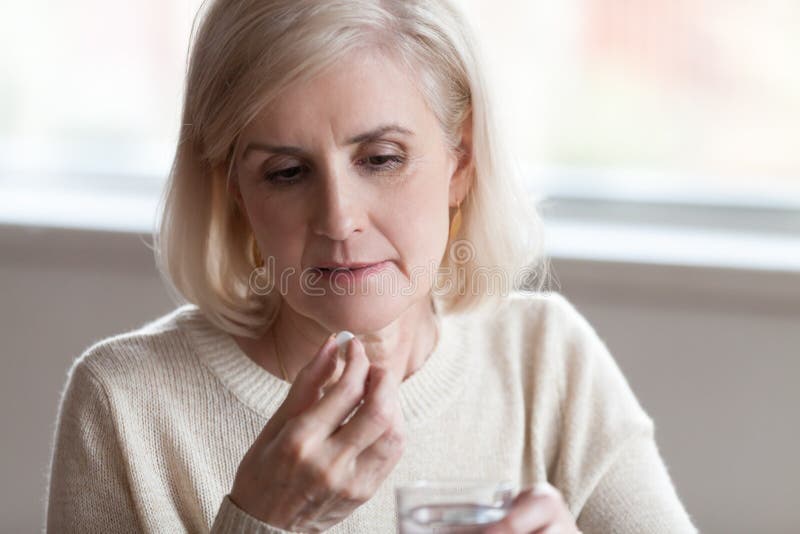 Close up of sick mature woman holding glass of still water and medical pill suffering from migraine headache feeling unhealthy and unwell. Many diseases are associated with age-related changes concept. Close up of sick mature woman holding glass of still water and medical pill suffering from migraine headache feeling unhealthy and unwell. Many diseases are associated with age-related changes concept