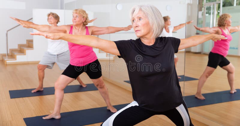 Active aged woman doing yoga with group in fitness studio, standing in lunging asana Virabhadrasana . Active aged woman doing yoga with group in fitness studio, standing in lunging asana Virabhadrasana .