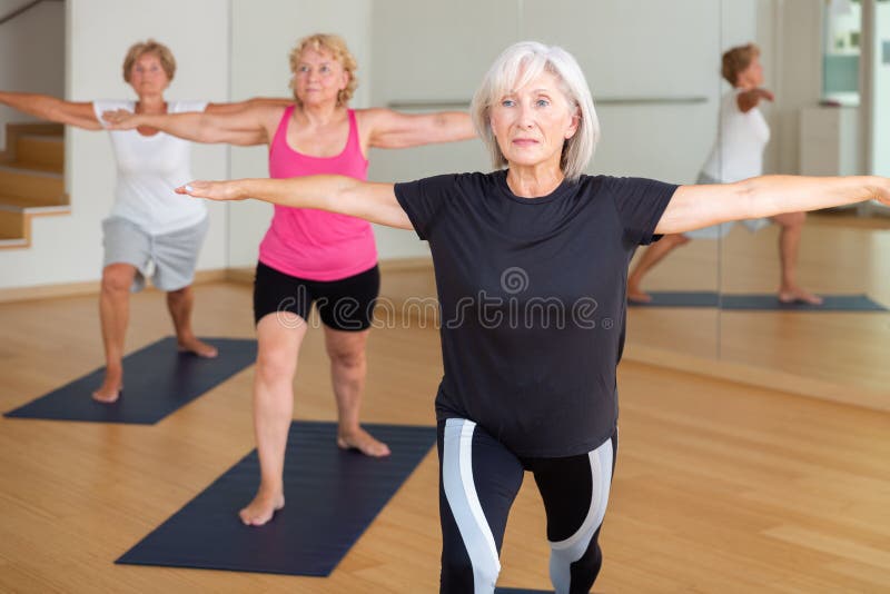 Active aged woman doing yoga with group in fitness studio, standing in lunging asana Virabhadrasana . Active aged woman doing yoga with group in fitness studio, standing in lunging asana Virabhadrasana .