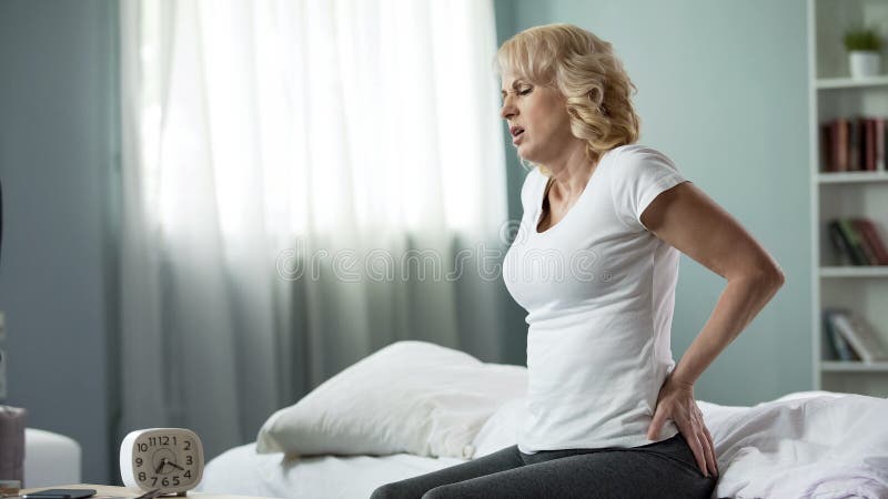 Aged woman suffering from low back pain, sitting on bed, spinal disk herniation, stock photo
