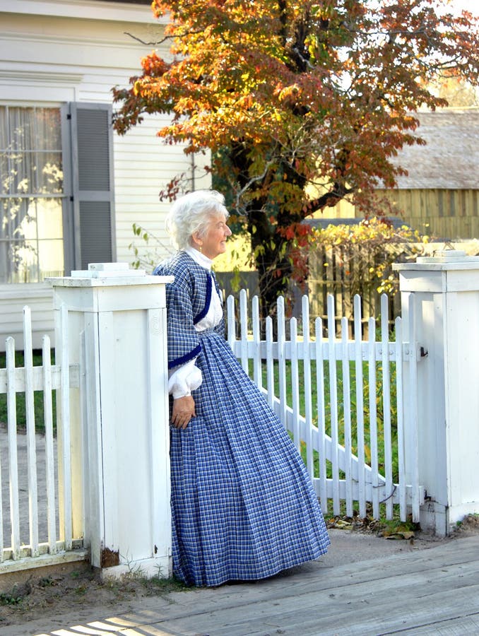 Re-enactment volunteer watches at gate for her returning war hero son or husband. White wooden fence and sidewalk. Re-enactment volunteer watches at gate for her returning war hero son or husband. White wooden fence and sidewalk.