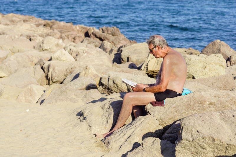 Agradável, França - 5 de agosto de 2017: opinião o homem que senta-se nas rochas que relaxam e lidas um livro na praia fotografia de stock royalty free Agradável, França - 5 de agosto de 2017: opinião o homem que senta-se nas rochas que relaxam e lidas um livro na praia fotografia de stock royalty free