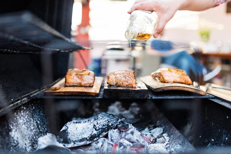 Image of a grill with salmon filet on wooden plank. The cooks is pouring aromatic alcohol on the fish for a stronger flavor. Image of a grill with salmon filet on wooden plank. The cooks is pouring aromatic alcohol on the fish for a stronger flavor