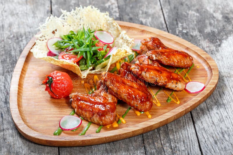 Ailes de poulet frit avec de la salade fraîche, les légumes grillés et la sauce à BBQ sur la planche à découper sur le fond en bo photographie stock libre de droits Ailes de poulet frit avec de la salade fraîche, les légumes grillés et la sauce à BBQ sur la planche à découper sur le fond en bo photographie stock libre de droits