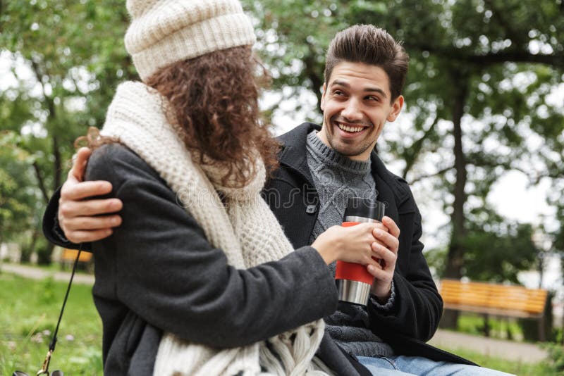 Aire libre de consumición del té o del café de los pares cariñosos jovenes felices en el parque que habla con uno a imagen de archivo libre de regalías Aire libre de consumición del té o del café de los pares cariñosos jovenes felices en el parque que habla con uno a imagen de archivo libre de regalías
