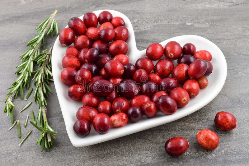 Heart healthy red Cranberries sit merrily on a heart shaped plate. Fresh organic rosemary graces the dark marble background to create a festive holiday or heart health concept. Heart healthy red Cranberries sit merrily on a heart shaped plate. Fresh organic rosemary graces the dark marble background to create a festive holiday or heart health concept