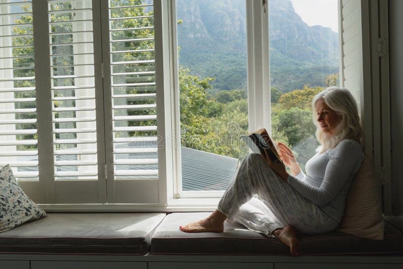 Side view of active senior Caucasian woman reading a book on window seat at home. Side view of active senior Caucasian woman reading a book on window seat at home