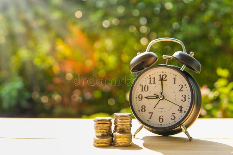 Alarm clock and coins on the wooden table in the sunset, money concept.