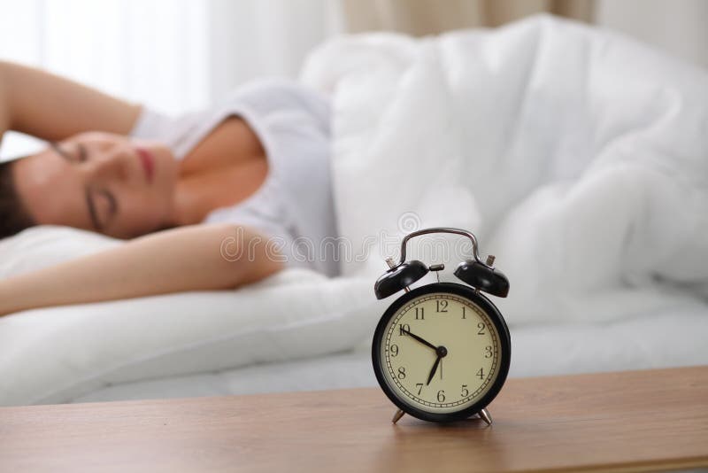 Alarm clock standing on bedside table has already rung early morning to wake up woman in bed sleeping in background