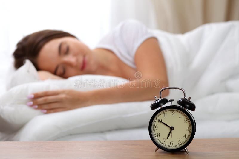 Alarm clock standing on bedside table has already rung early morning to wake up woman in bed sleeping in background