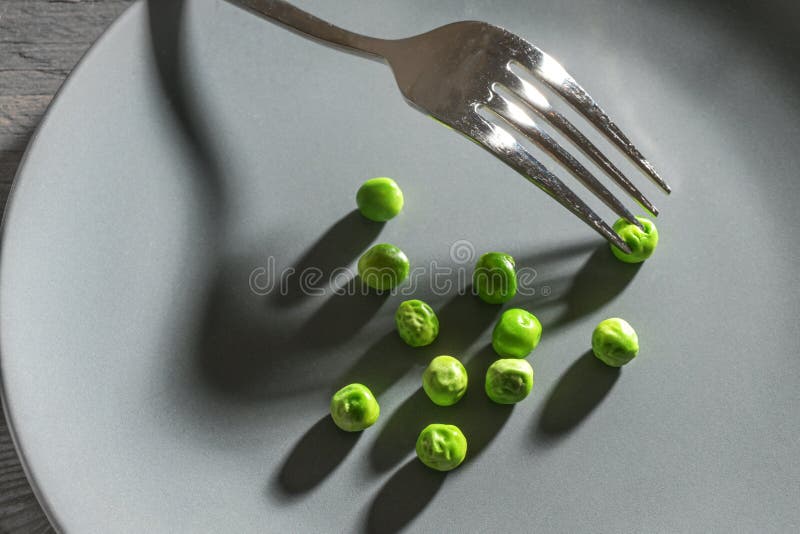 Some green peas and a fork with shadows on a gray plate, meager diet meal after the resolution to slim down, copy space, selected focus. Some green peas and a fork with shadows on a gray plate, meager diet meal after the resolution to slim down, copy space, selected focus