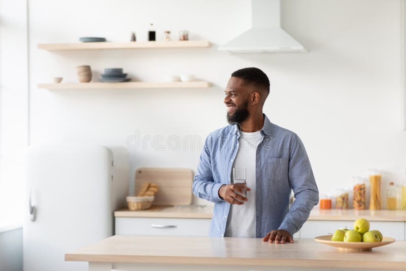 Alegre joven negro barbudo con vaso de agua limpia en la cocina interior mira a la ventana foto de archivo