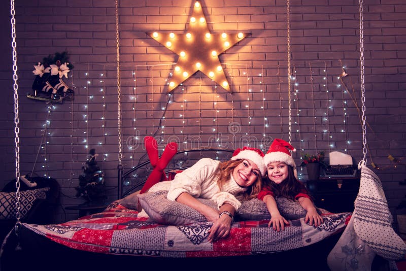 Alegre linda madre e hija con sombreros de navidad contra la iluminación en forma de estrella en una cama suspendida con cadenas imagenes de archivo