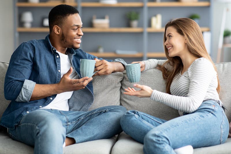 Alegre pareja mixta relajarse en el sofá en casa con café y charlar imagen de archivo