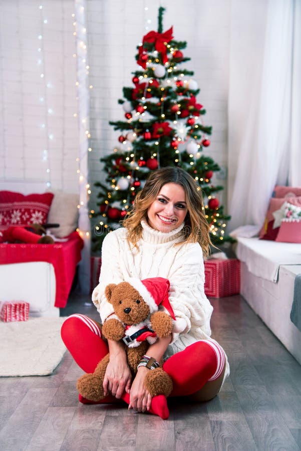 Alegre y bonita madre con suéter de punto blanco y leggings rojas con un juguete de oso de peluche de navidad de navidad con fondo foto de archivo libre de regalías