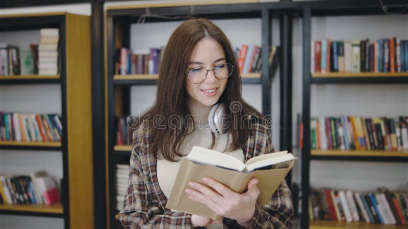 Alla giovane donna piace leggere un libro in una biblioteca con scaffali pieni di libri mentre indossa le cuffie video d archivio