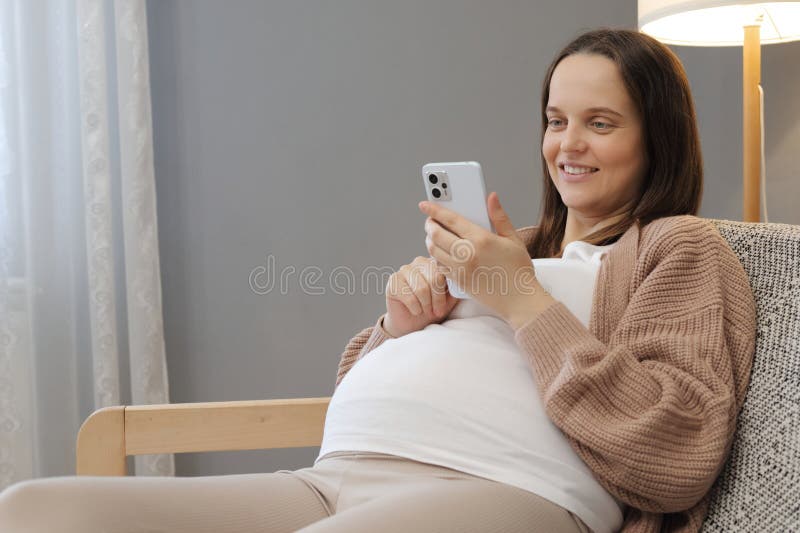 Allegra e soddisfatta giovane donna incinta che usa uno smartphone per navigare su pagine web leggendo informazioni su forum per immagini stock libere da diritti