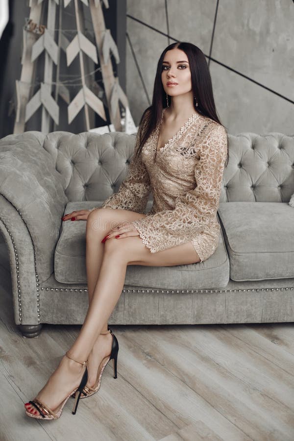 Full length of gorgeous model with brunette hair and make up wearing luxurious beige laced dress with long sleeves and high heels. She is sitting on expensive grey couch and looking at camera. Full length of gorgeous model with brunette hair and make up wearing luxurious beige laced dress with long sleeves and high heels. She is sitting on expensive grey couch and looking at camera.