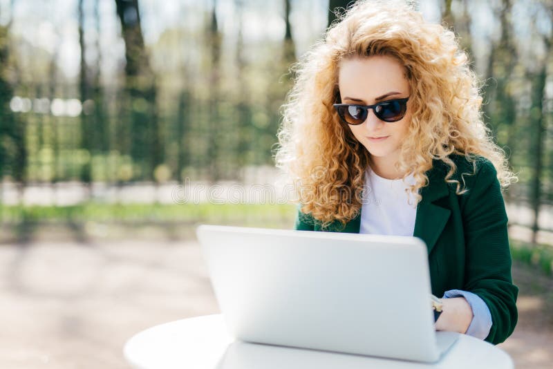Allvarlig kvinnlig student som har fluffigt blont hår som bär det gröna omslaget och solglasögon som sitter göra utomhus hennes d arkivbilder Allvarlig kvinnlig student som har fluffigt blont hår som bär det gröna omslaget och solglasögon som sitter göra utomhus hennes d arkivbilder