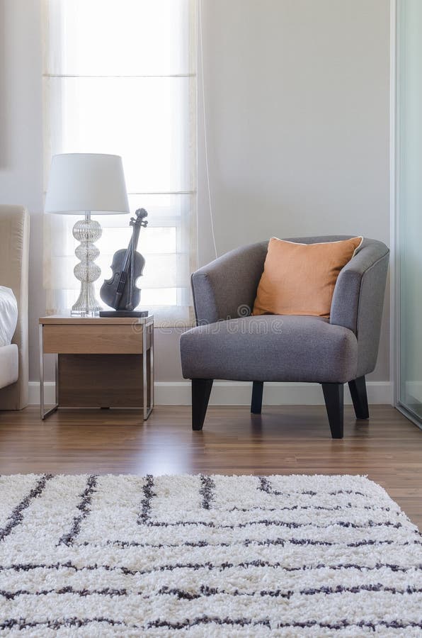 Orange pillow on modern grey chair with bedside table and white lamp in bedroom at home. Orange pillow on modern grey chair with bedside table and white lamp in bedroom at home