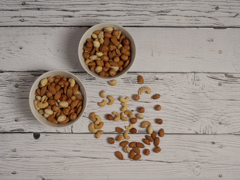 Almonds, hazelnuts, cashews and pistachios nuts in two bowls on a white wooden table top view. Some nuts are scattered on the table