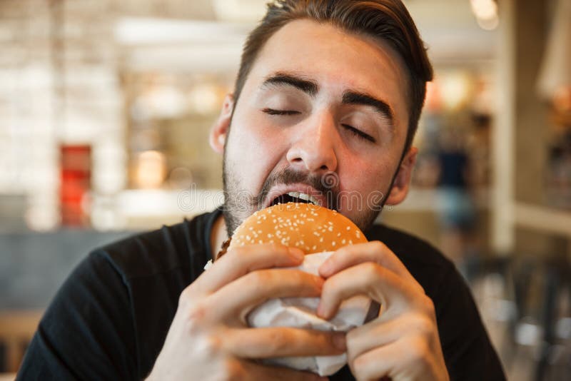 Almuerzo en el caf? Mordeduras una hamburguesa con los ojos cerrados fotografía de archivo libre de regalías Almuerzo en el caf? Mordeduras una hamburguesa con los ojos cerrados fotografía de archivo libre de regalías