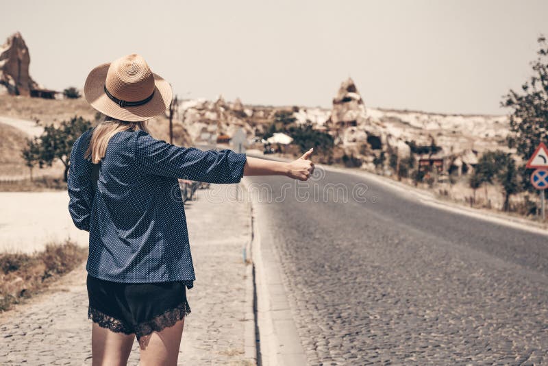 Alone hitchhiker girl in straw hat stands on the roadside. Arm with the thumb pointing upward. Mountains on the royalty free stock photo Alone hitchhiker girl in straw hat stands on the roadside. Arm with the thumb pointing upward. Mountains on the royalty free stock photo