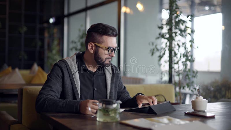 Alone young man is resting alone in cafe in daytime, using tablet and sipping hot drink tea from cup, browsing by wifi. Hot cafe video