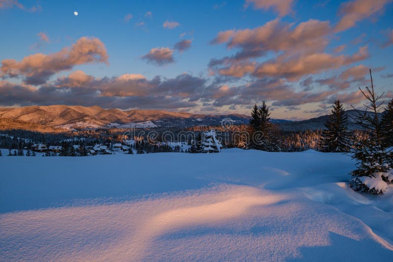 Alpine village outskirts in last evening sunset sun light. Winter snowy hills and fir trees. Picturesque clouds and Moon in dusk stock image