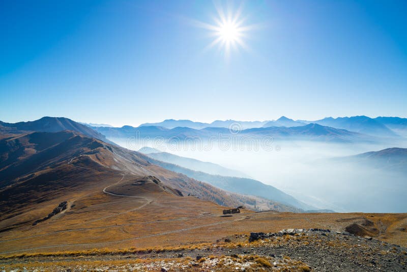 The Alps in backlight from mountain top. Autumn view with bright sunburst and clear blue sky. Fog and mist in the valley below.