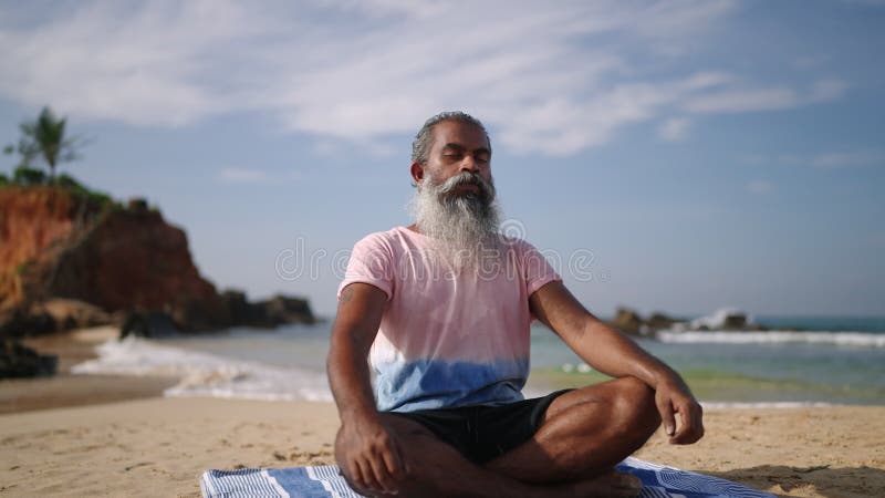 Alter Mann meditieren und sitzen in lotus posieren zum Geräusch der Wellen auf dem paradiesischen Inselstrand am Meer. schwarzes M stock video footage